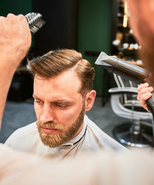 Experienced barber using hairdryer with brush to style hair.