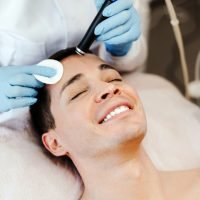 Profession cosmetologist making vacuum facial skin cleaning for smiling man in beauty salon